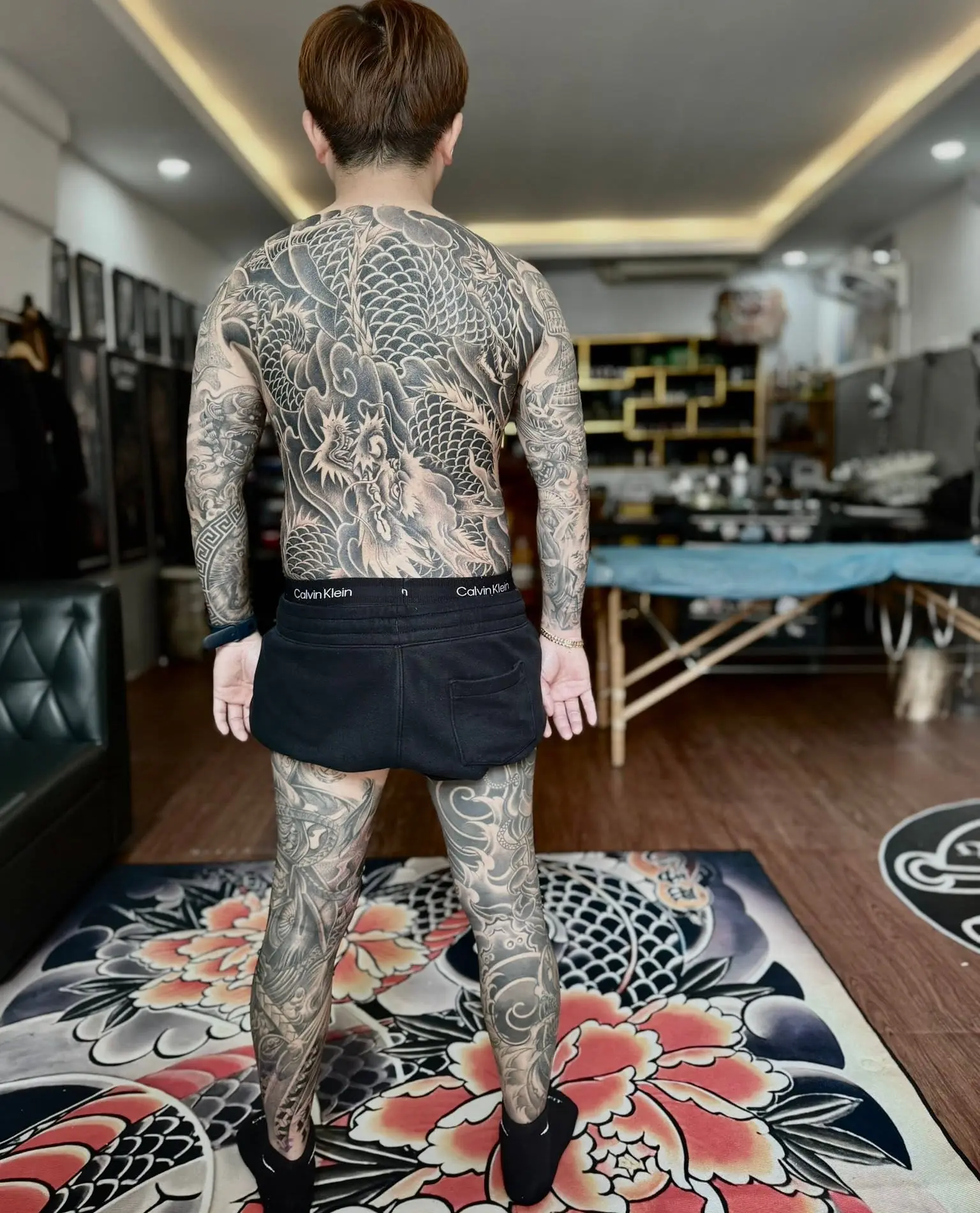 A client standing in the studio wearing a full Japanese bodysuit — koi, dragon and peony work covering back, arms and legs — tattooed by Meng