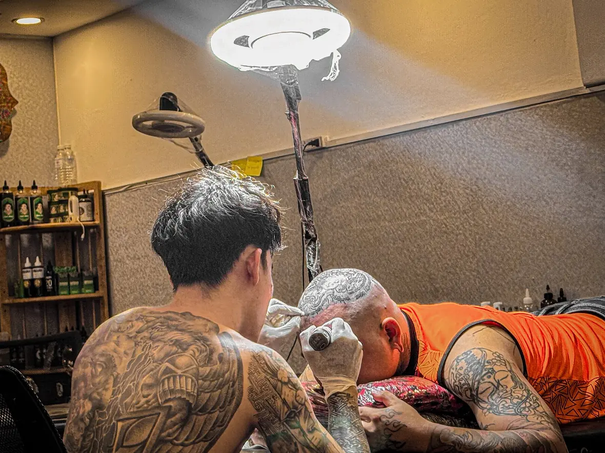 Meng leaning in over a client under the studio lamp at 55 Tattoo