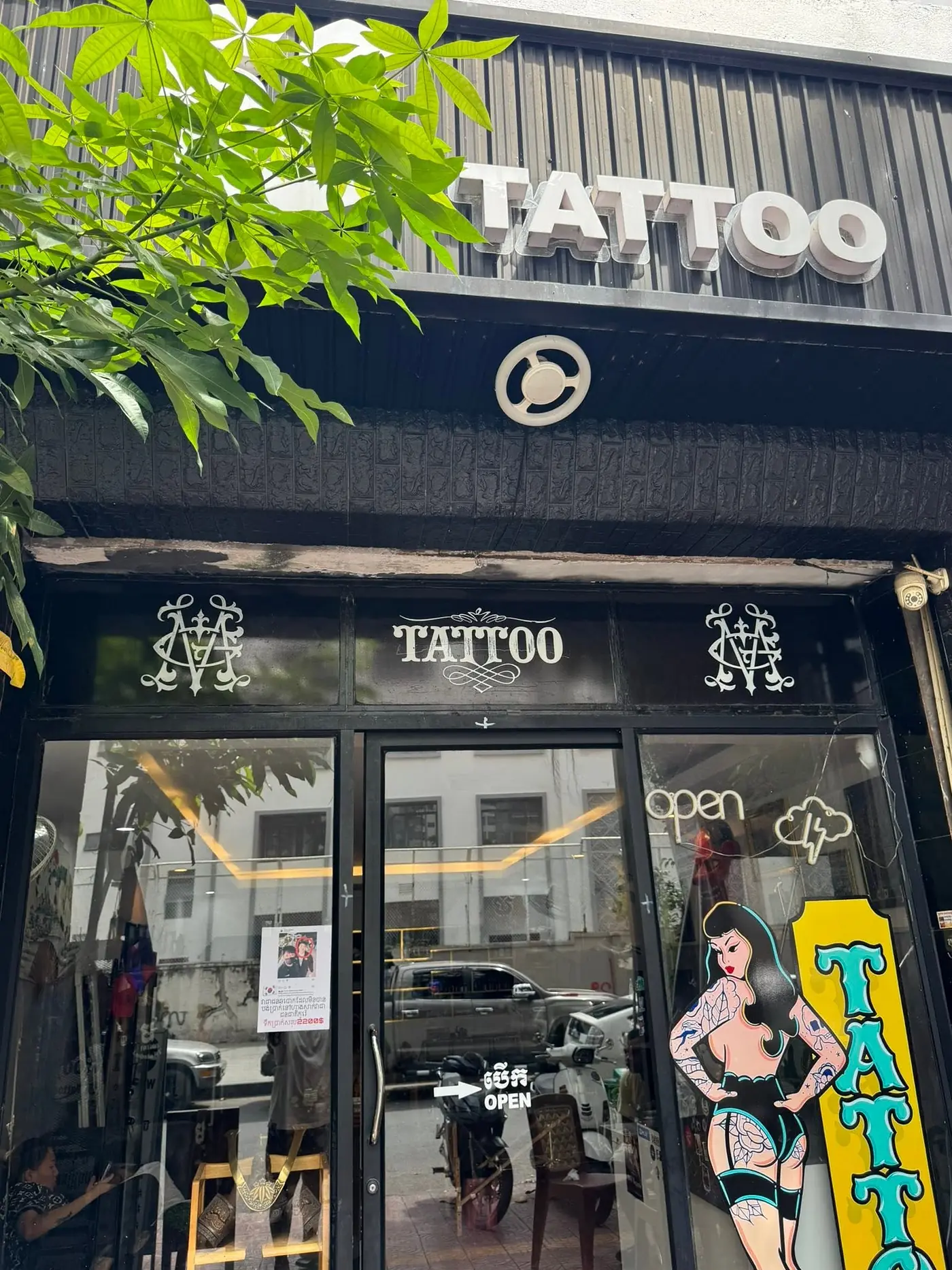 The 55 Tattoo shopfront on Street 90 — Meng's ornate monogram painted on the glass either side of the word TATTOO, under black-painted Khmer temple relief